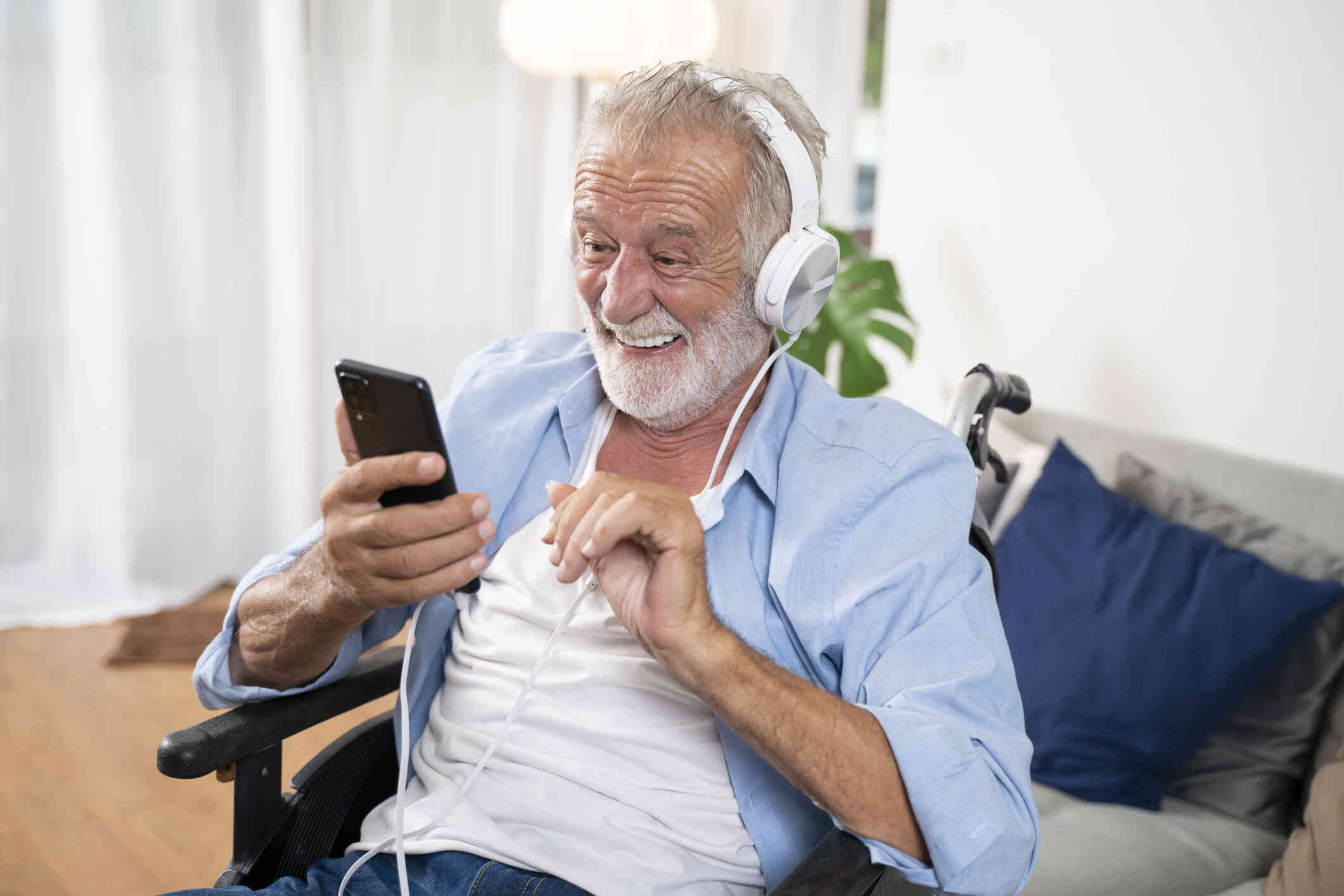 An elderly man in a wheelchair sits in his living room and smiles brightly at the cellphone in his hand while wearing white headphones.